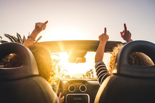 Couple of woman friends traveling and driving having a lot of fun dancing in the car with opened roof and summer vacation sunset ocean in front