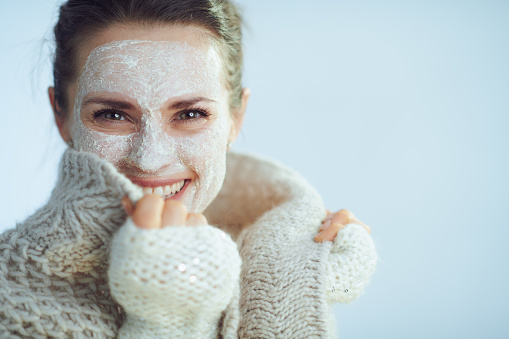 happy modern woman in roll neck sweater and cardigan with white facial mask as part of winter skin care covering in clothes isolated on winter light blue background.