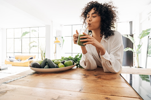 Young african american woman drinking green juice with reusable bamboo straw in loft apartment. Home concept. Healthy lifestyle concept.