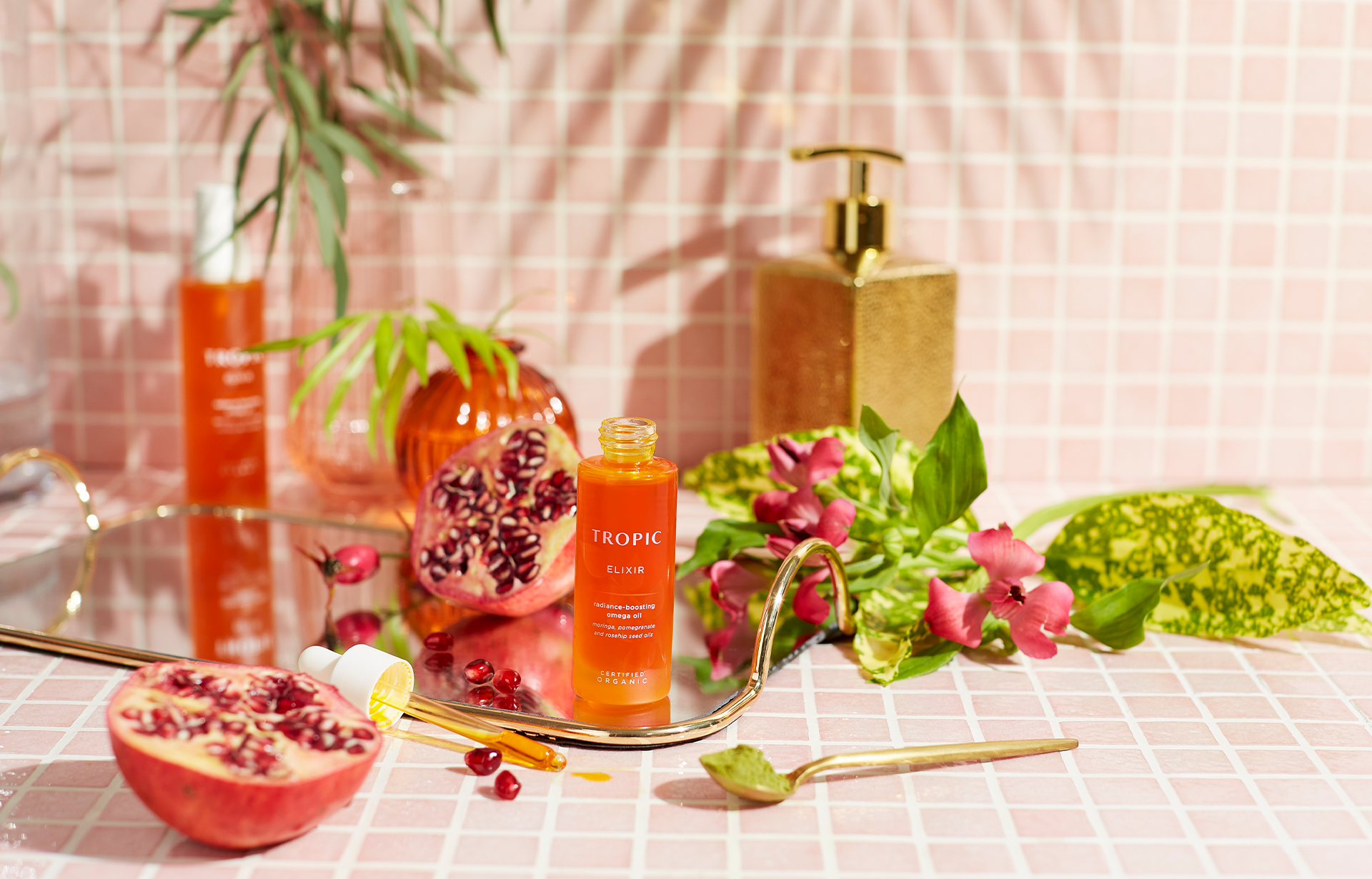 Light pink tiled background with leaves and pomengrante cut in half. In the middl is Elixir facial oil with the pipette to the left.