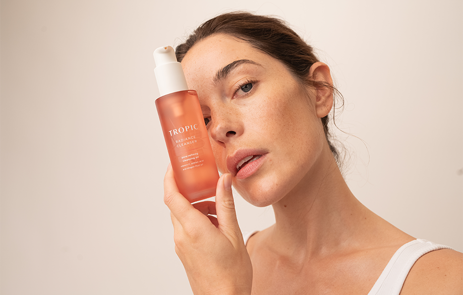 The image shows a woman holding a bottle of Tropic Radiance Cleanser next to her face. The bottle is a light pink with a white pump dispenser. The woman has clear, glowing skin and is wearing a white tank top with her hair pulled back, creating a clean and fresh aesthetic.