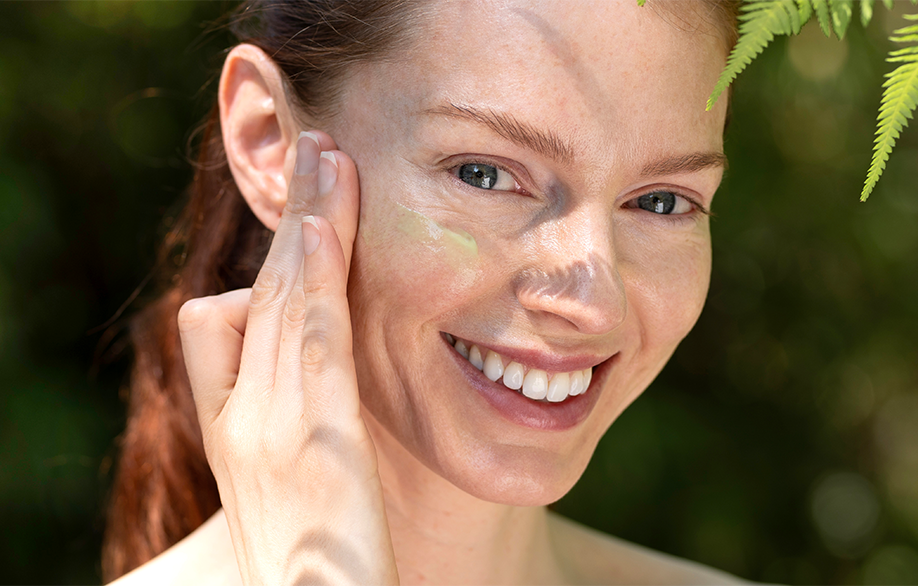 Woman applies Skin Re-Leaf moisturiser to her face and her hand rests beside it as she smiles. The background is green with leaves just above and infront of her face.